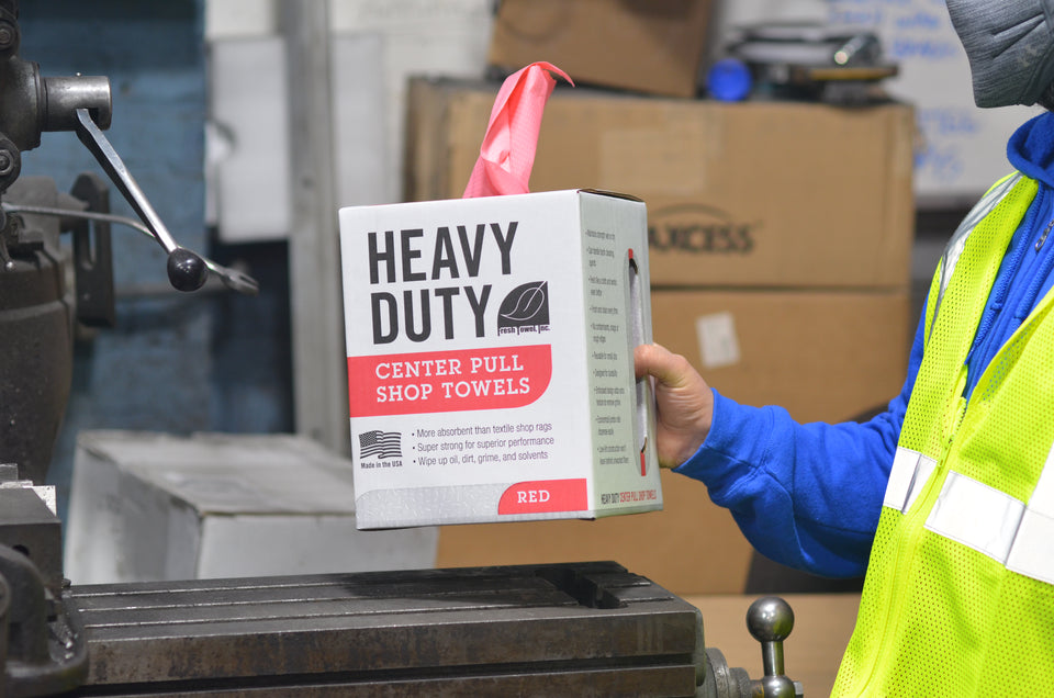 Person holding box of Red Heavy Duty Center Pull Shop Towels by perforated punch-out handle at corner of box in a mechanic shop with a safety vest and mask.