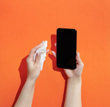 Load image into Gallery viewer, Orange background with hands wiping smartphone with a wet wipe.